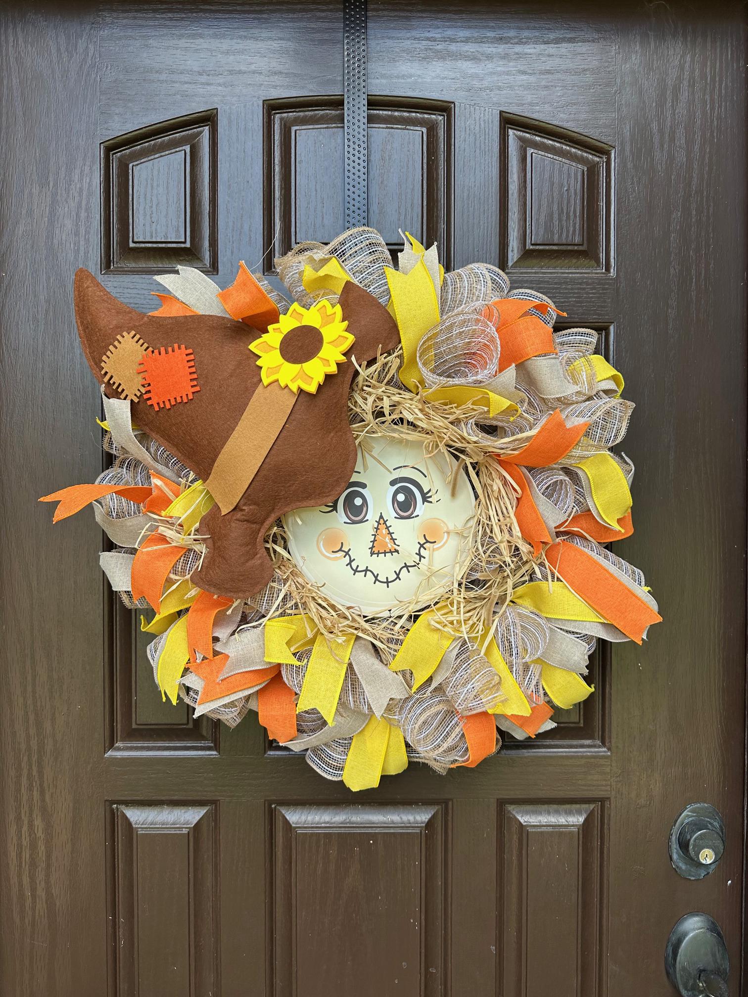 A scarecrow face at the center of a ribbon wreath with a brown hat.