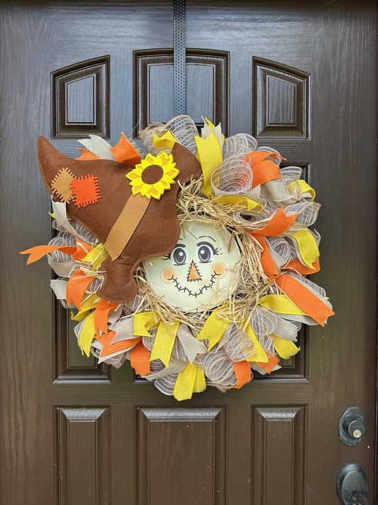 A scarecrow face at the center of a ribbon wreath with a brown hat.